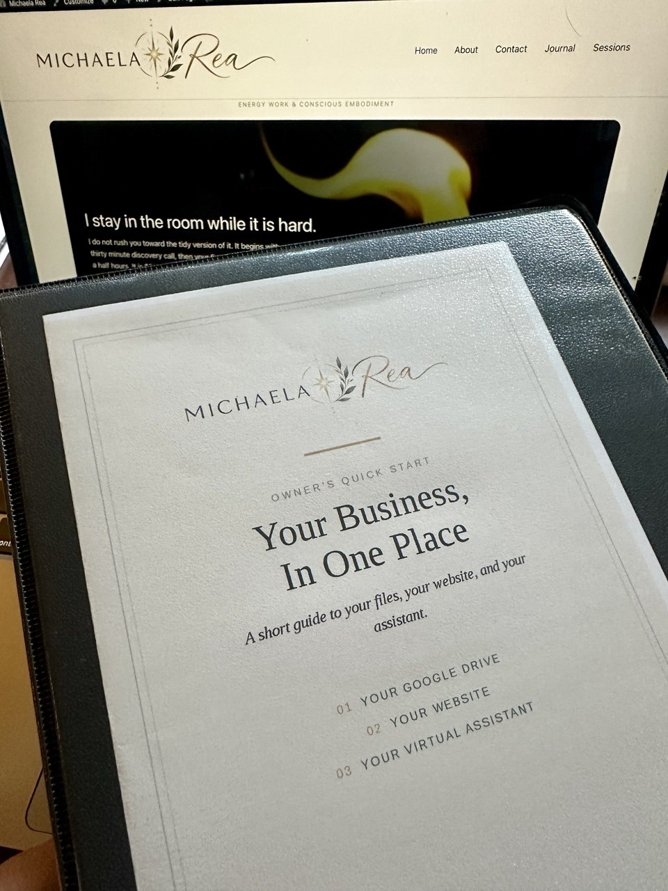A printed owner's quick start binder standing in front of a laptop showing the Michaela Rea website